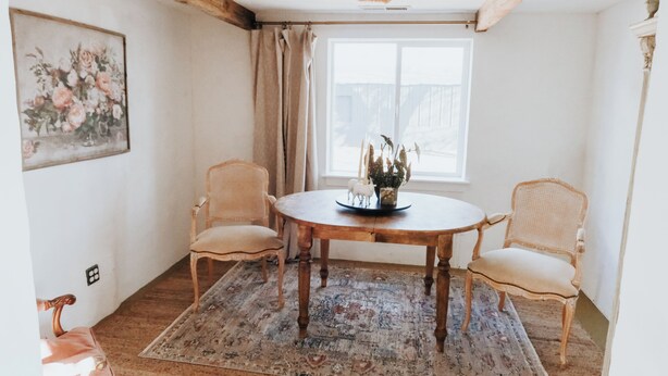 A dining area with a round wooden table, two wicker chairs, a floral painting on the wall, a window with curtains, and a chandelier.