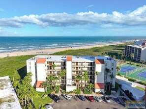 Ein Hotel direkt am Strand mit mehreren Etagen, einem Parkplatz und einem Tennisplatz.