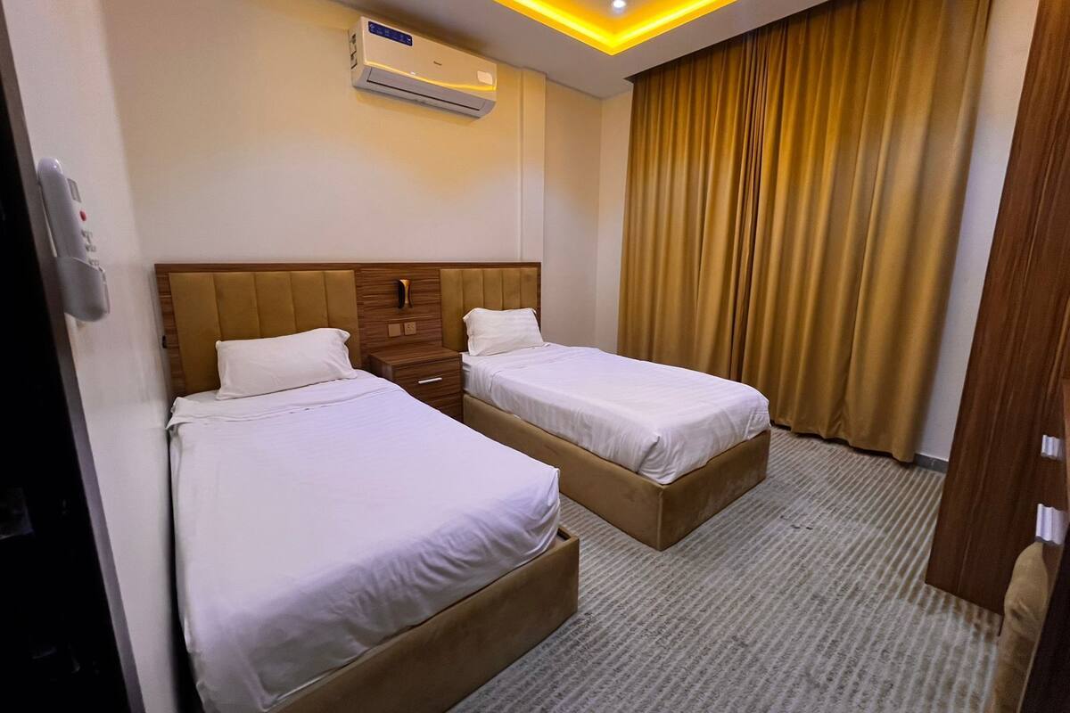 Een hotelkamer met twee bedden, een houten hoofdbord, een nachtkastje, een airconditioning en een groot raam met gordijnen.