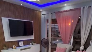 A modern living room with a flat-screen TV, a white sofa with pink cushions, a ceiling fan, a large window with curtains, and a staircase visible through the window. - Elegant & Comfortable Apartment with Private Balcony (Santo Domingo Este)