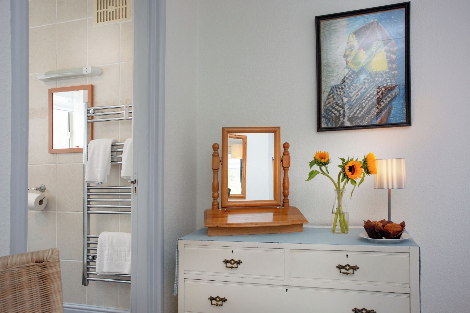 A bathroom with a white vanity, a wooden-framed mirror, a vase with sunflowers, and a framed painting on the wall.