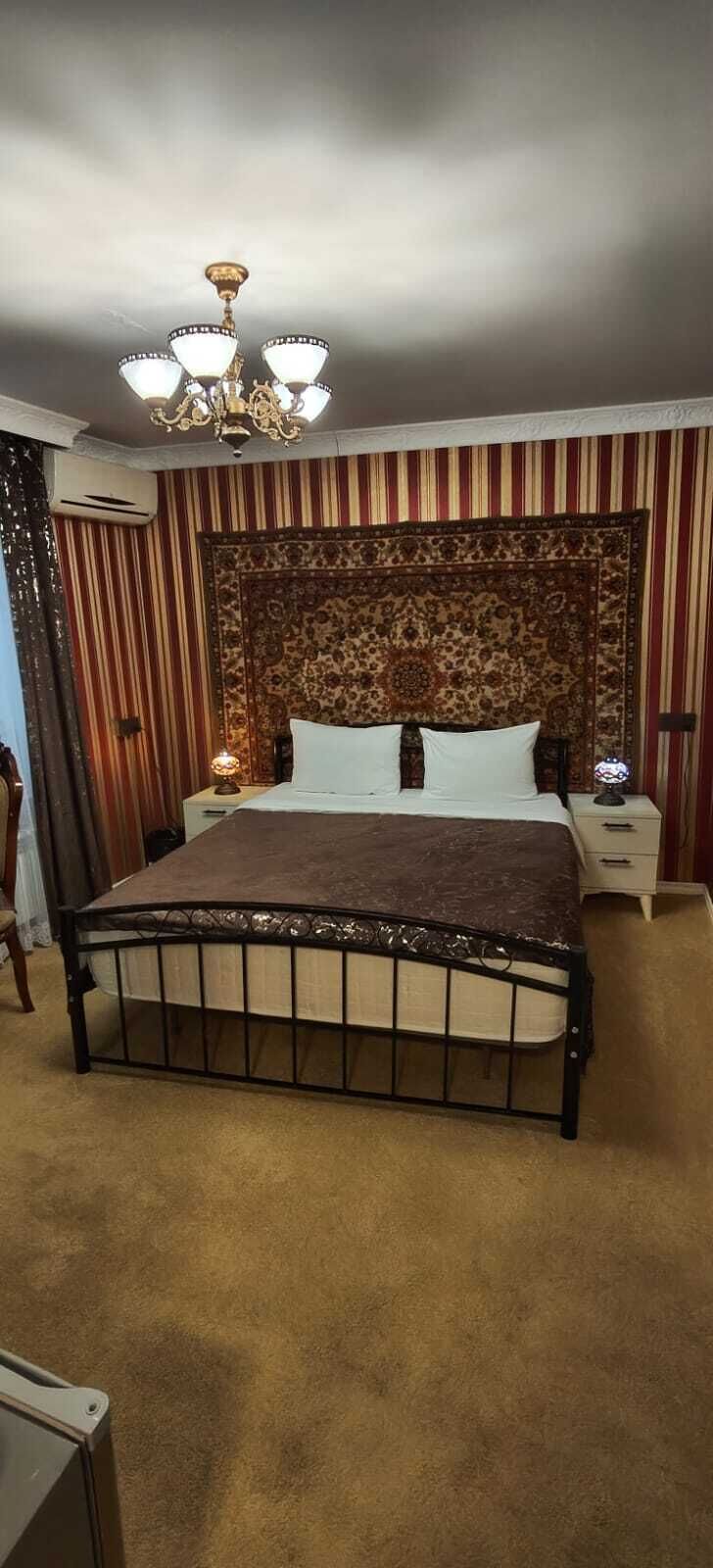 A bed with a headboard, bedside tables, and a chandelier in a room with striped walls and a carpeted floor.
