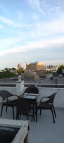 A rooftop terrace with wicker chairs and a table, overlooking a cityscape with a prominent building and a dome structure.