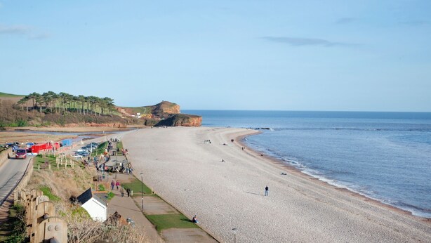 Ein Strand mit Kieselsteinen, eine Klippe mit Bäumen und eine Straße mit Fahrzeugen und Fußgängern.