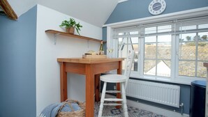 Ein Zimmer mit einem Holztisch, einem weißen Stuhl, einem Blumentopf, einer Uhr und einem Fenster mit Jalousien.