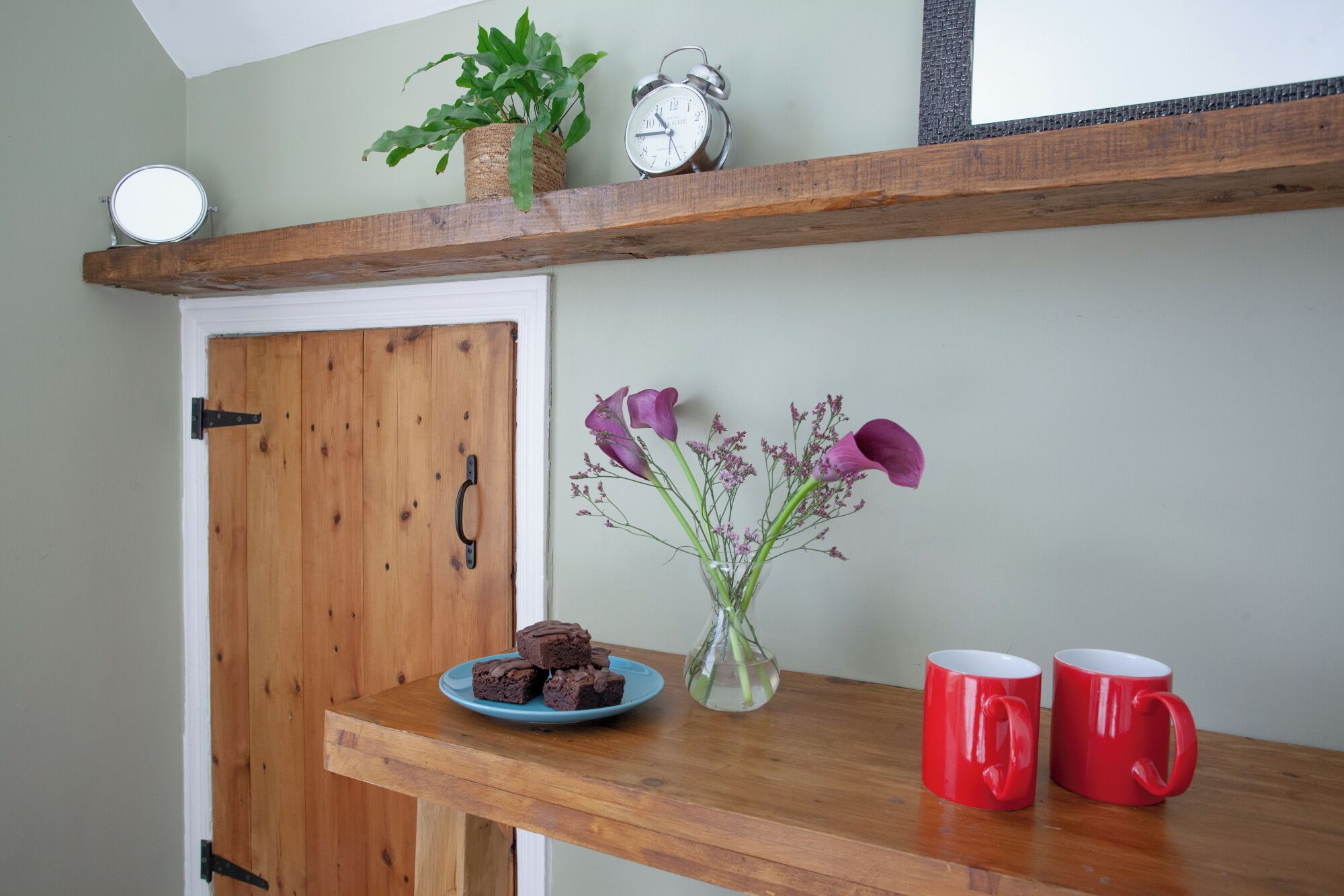 A wooden door with a handle, a shelf with a clock, a vase with flowers, and two red mugs on a wooden table.
