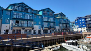 Une rangée de bâtiments bleus à plusieurs étages, dotés de balcons, un quai avec des bateaux et un ciel bleu dégagé.
