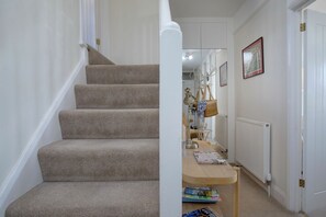 Una escalera con alfombra que lleva a una habitación con radiador, una mesa con libros y un perchero con cesta.