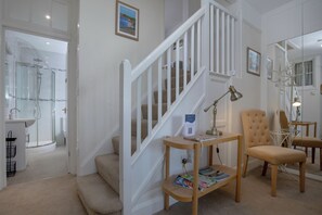 Una escalera con barandilla blanca que conduce a una plataforma, una mesa de madera con revistas y un sillón color beige.