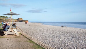 A pebbled beach with picnic tables and umbrellas, a cliff in the background, and people walking and sitting. - Budleigh Breeze - Three Bedroom Apartment, Sleeps 6 (Budleigh Salterton)