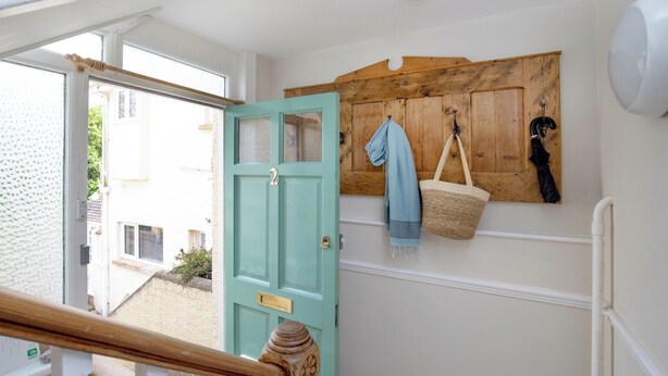 A staircase with a wooden handrail leading to a teal door. A wooden wall-mounted rack holds a blue towel and a woven basket. - Budleigh Breeze - Three Bedroom Apartment, Sleeps 6 (Budleigh Salterton)