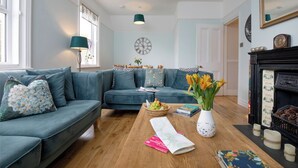A living room with a blue sofa set, a wooden coffee table with books and a vase of flowers, a fireplace, and a clock on the wall. - Budleigh Breeze - Three Bedroom Apartment, Sleeps 6 (Budleigh Salterton)