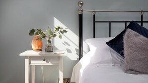 A neatly made bed with white linens and a dark blue pillow, a white bedside table with a vase of green plants, and a black metal headboard. - Budleigh Breeze - Three Bedroom Apartment, Sleeps 6 (Budleigh Salterton)