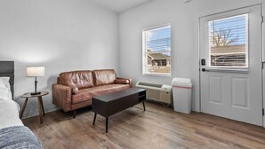 Een kleine, goed verlichte kamer met een bruine leren bank, een klein rond tafeltje met een lamp, een donker houten salontafel, een witte deur en een raam met jaloezieën.
