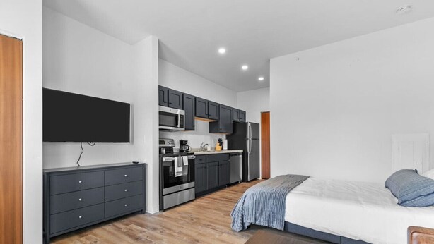 Een moderne, compacte slaapkamer met een groot bed en een houten nachtkastje. Grenzend aan de kamer bevindt zich een kitchenette met donkere kastjes, een magnetron en een koelkast.