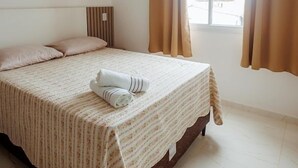 A neatly made bed with a striped bedspread, a pillow, and a rolled towel on the bed. There is a window with curtains, a ceiling light, and a headboard with vertical lines. - Apto c Garagem e Ar Condicionado na Praia do Sul (Ilhéus)