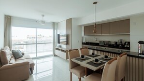 A modern apartment with a dining area, kitchen, and a large window offering a city view. - Apto c Garagem e Ar Condicionado na Praia do Sul (Ilhéus)
