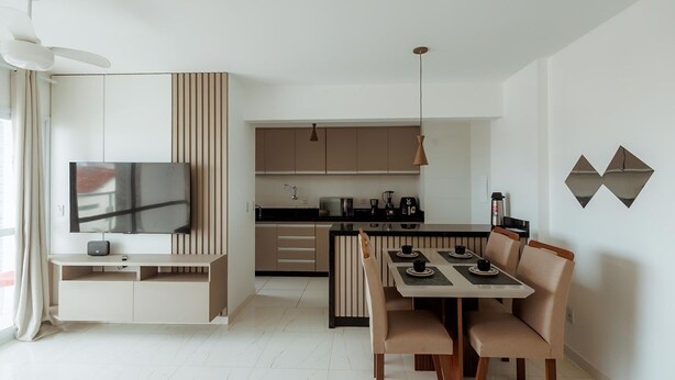 A modern living room with a flat-screen TV, a dining area with a table and chairs, and a kitchen with a black countertop and wooden cabinets. - Apto c Garagem e Ar Condicionado na Praia do Sul (Ilhéus)