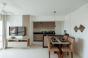 Ein modernes Wohnzimmer mit Flachbildfernseher, einem Essbereich mit Tisch und Stühlen sowie einer Küche mit schwarzer Arbeitsplatte und Holzschränken.