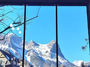 Blick auf schneebedeckte Berge durch ein Fenster mit kahlen Ästen.