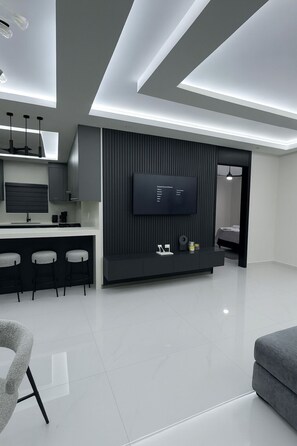 Un salon moderne doté d’un téléviseur à écran plat fixé sur un mur foncé, d’un élégant coin bar avec des tabourets et d’un design minimaliste.