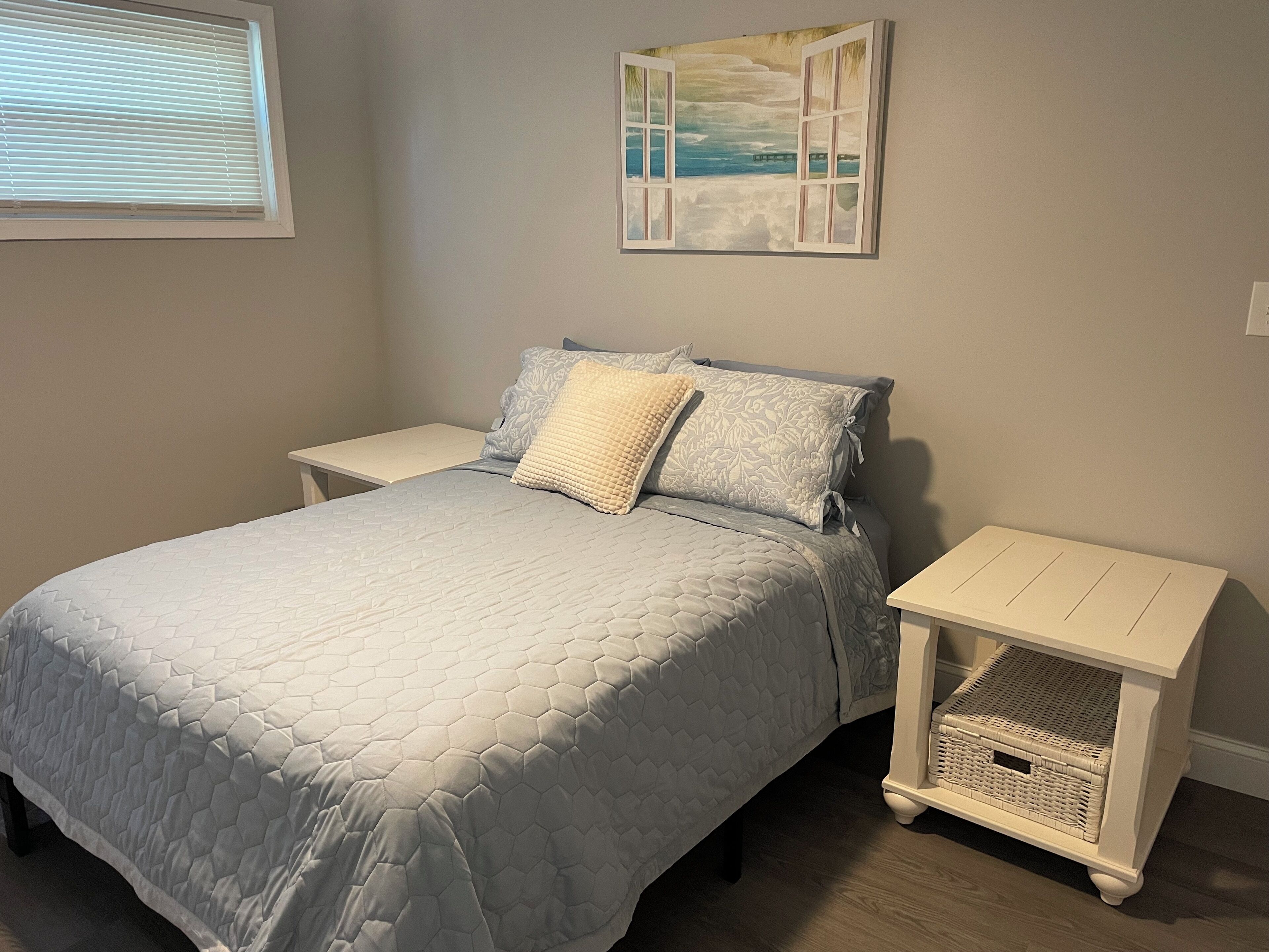 A bedroom with a bed, two nightstands, a wall-mounted painting, and a wicker basket on one nightstand.