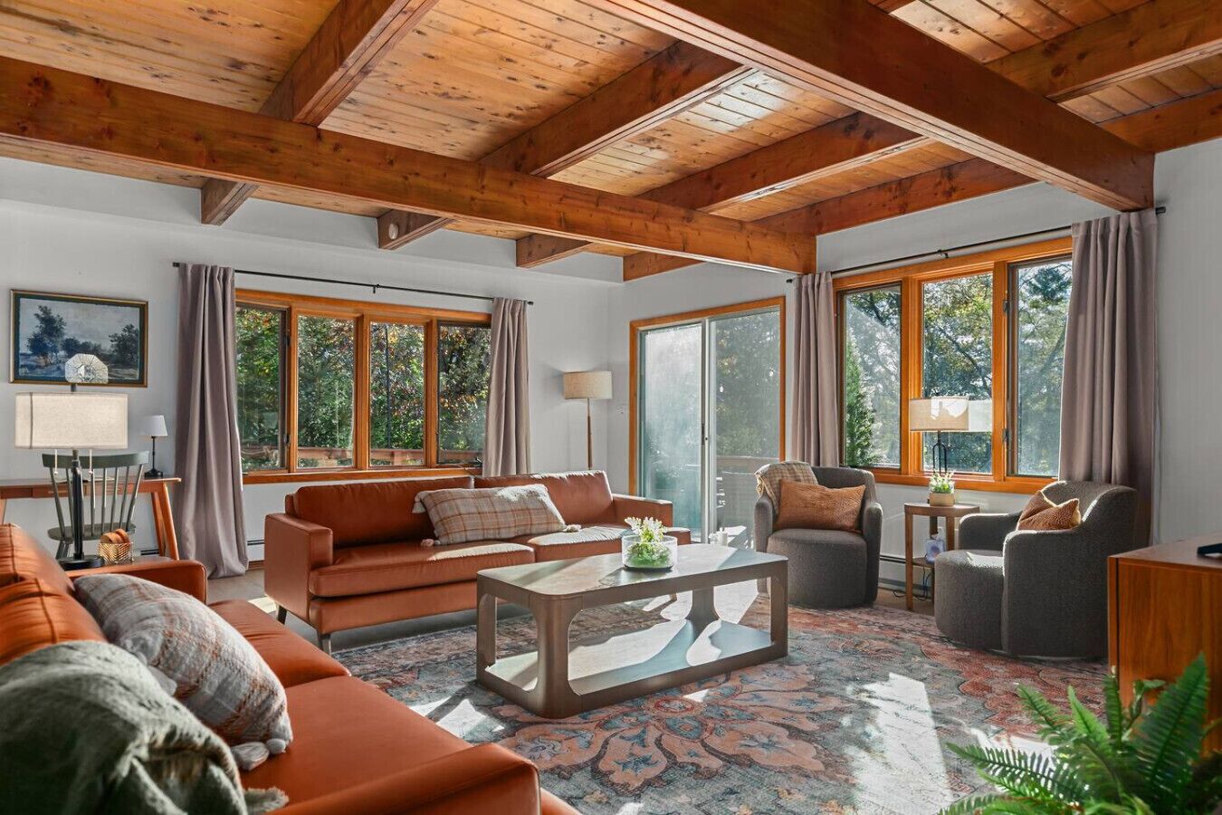 Un salon spacieux avec un haut plafond en bois, de grandes fenêtres, un ensemble de canapés, une table basse et une table d’appoint avec une lampe.