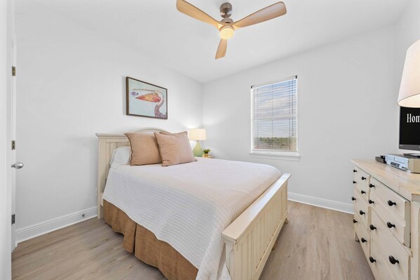 Quarto com cama, ventoinha no teto, janela com persianas, abajur e cômoda.