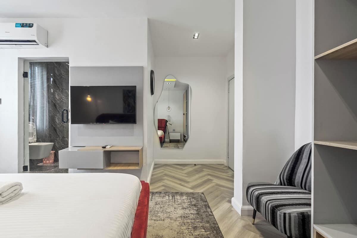A modern hotel room with a large bed, a flat-screen TV, a mirror, a wooden floor, and a striped chair.