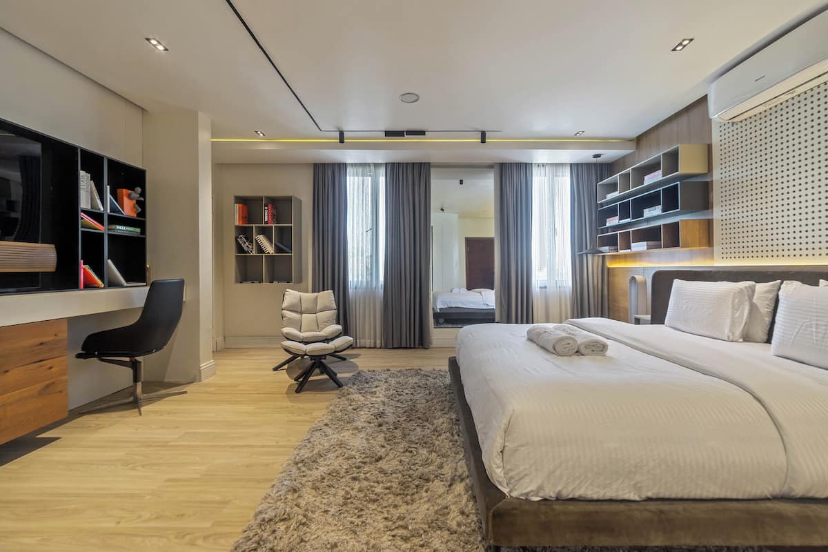 A modern hotel room with a large bed, a desk, a chair, and a bookshelf.