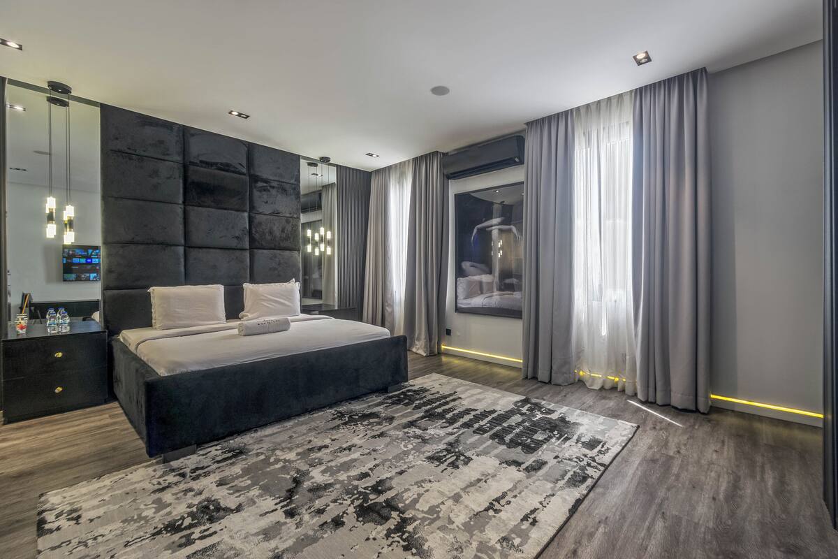 A modern bedroom with a large bed, a dark headboard, a large mirror, and a view of the city through the window.