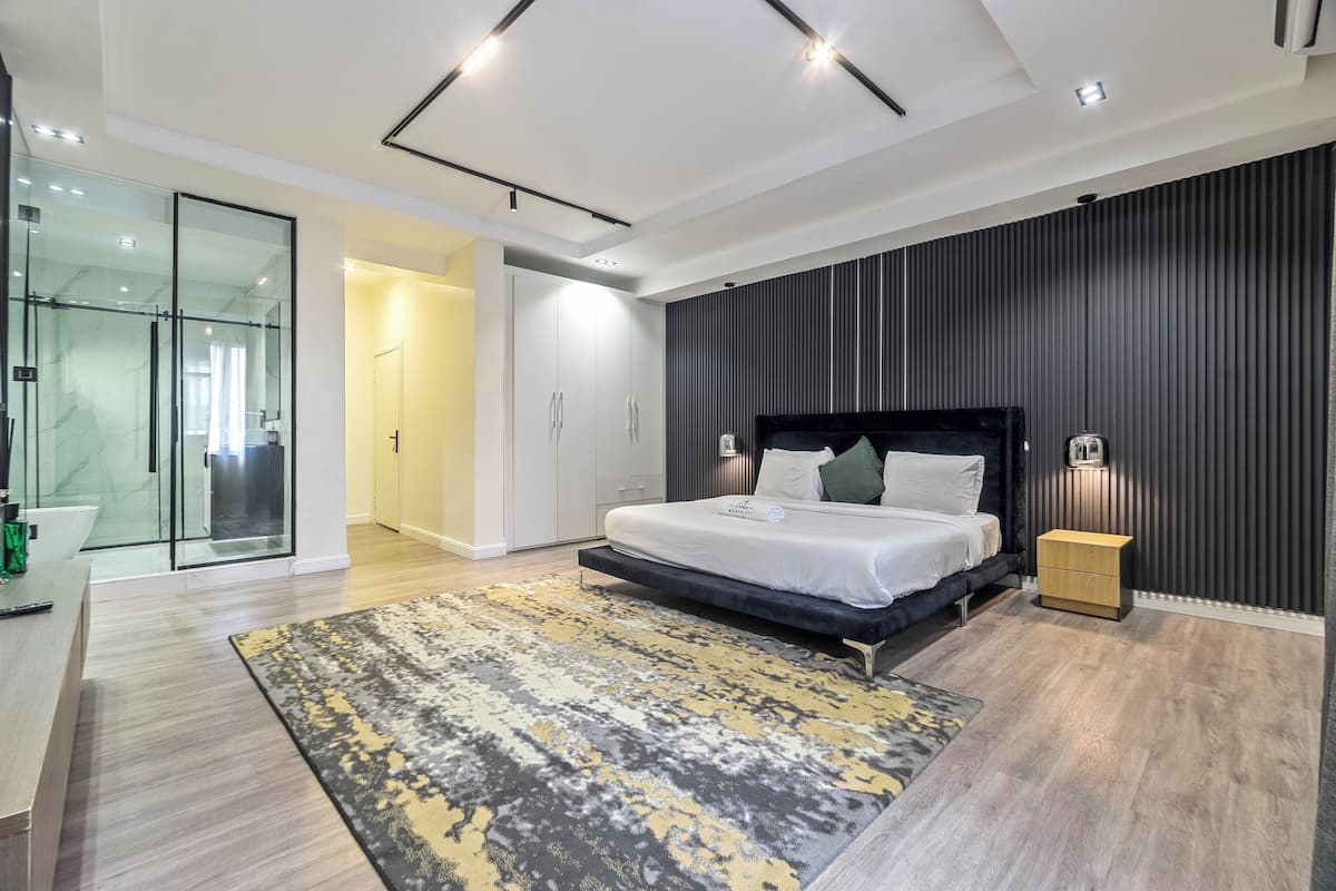 A modern bedroom with a large bed, bedside tables, a glass shower enclosure, and a dark wall paneling.
