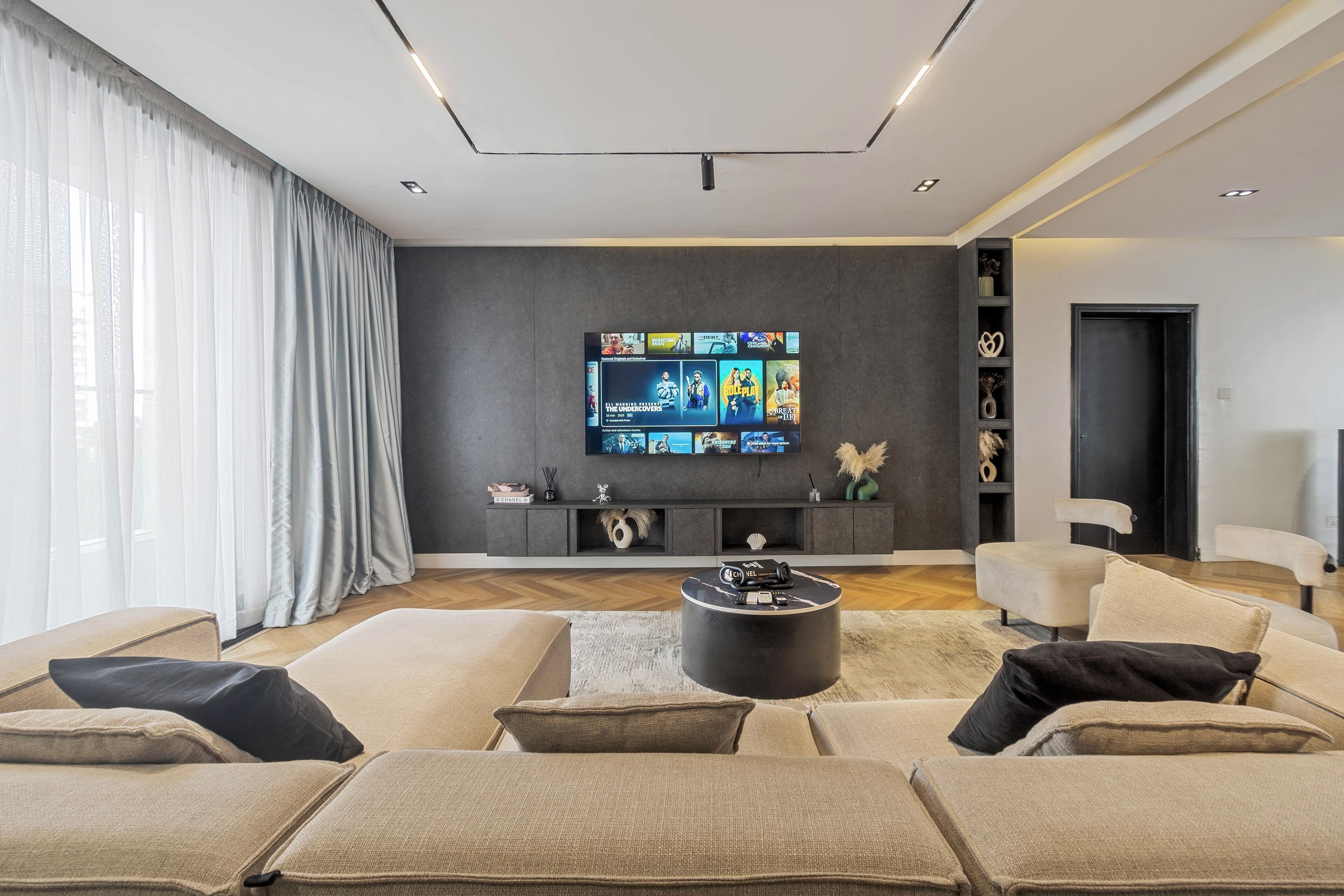 A modern living room with a large flat-screen TV mounted on a dark wall, a round coffee table, and a beige sectional sofa.