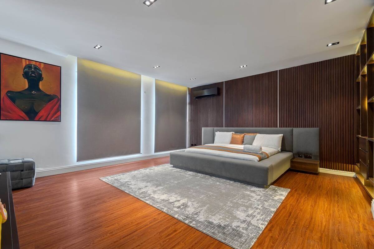 A modern bedroom with a large bed, a grey sofa, a wooden floor, and a large window.