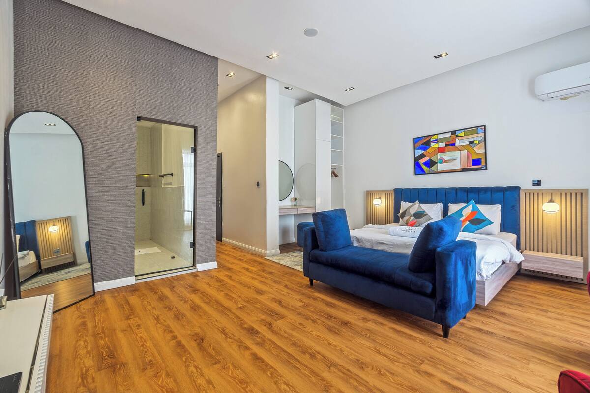 A modern hotel room with a wooden floor, a large bed with blue bedding, a blue sofa, a large mirror, and a glass shower enclosure.