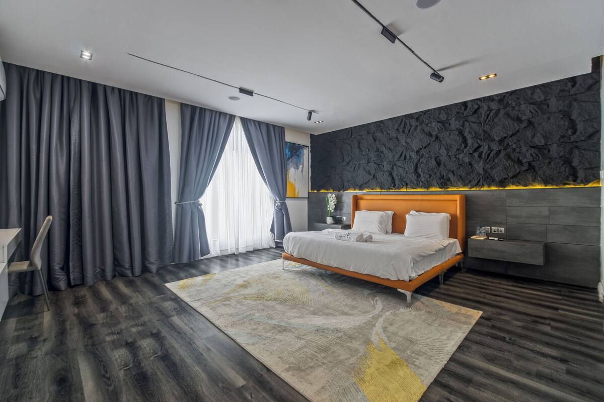 A modern bedroom with a large bed, dark wood flooring, a grey accent wall, and a large window with curtains.