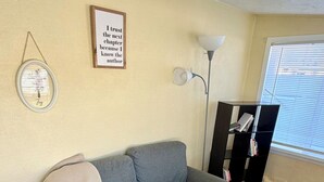 Ein Wohnzimmer mit grauem Sofa, einer Decke, einem gerahmten Zitat an der Wand, einer Stehlampe und einem Bücherregal.