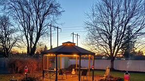 Ein Pavillon mit Dach, umgeben von einer grünen Rasenfläche, mit einem Feuerkorb in der Mitte.