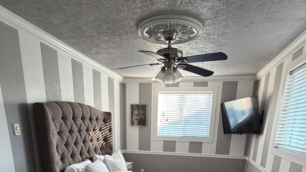 Ein Schlafzimmer mit einer gepolsterten Kopfteile, einem Bett mit weißer Bettwäsche, einem Deckenventilator, einem angebrachten Fernseher und einem gemusterten Teppich.