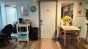 A compact living space with a kitchenette, a dining area with a table and chairs, a trash bin, a clock, and a door with a no-smoking sign.