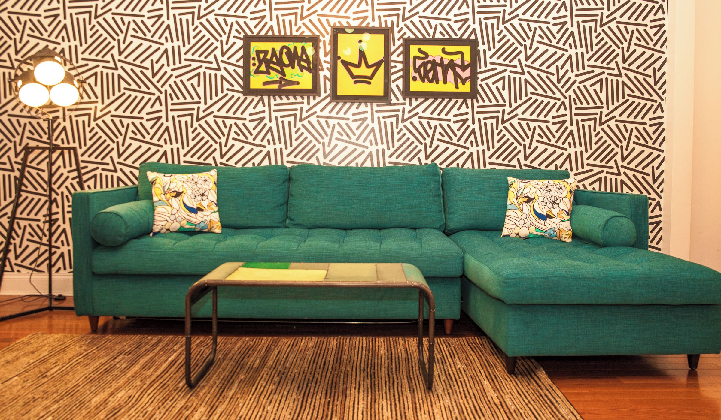A teal sectional sofa with floral cushions, a wooden coffee table, and a patterned wall with framed artwork.