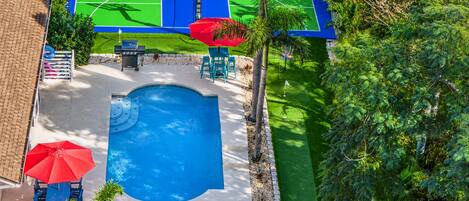 An aerial view of a backyard with a swimming pool, a basketball court, and a patio area with chairs and umbrellas.