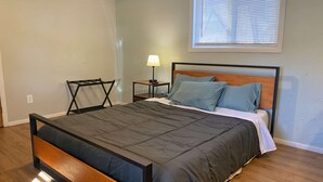Un dormitorio moderno con cabecera de cama de madera, ropa de cama gris, una mesita de noche con una lámpara y una ventana con persianas.