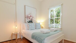 Ein ordentlich eingerichtetes Schlafzimmer mit Bett, Nachttischlampen, einem Fenster mit Jalousien und einem Wandbild.