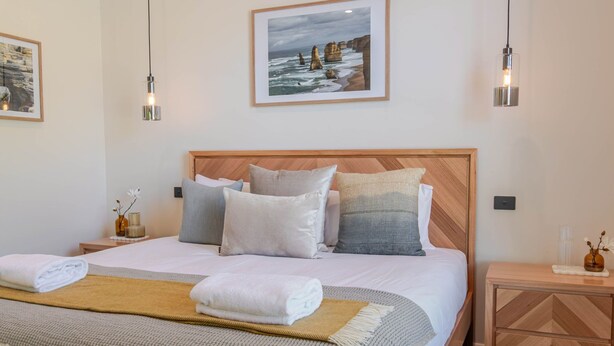 Een netjes opgemaakt bed met kussens en dekbed, een nachtkastje met een vaas, en een lijst met een foto aan de muur.