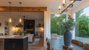 Une cuisine moderne dotée d’un îlot central, de poutres en bois au plafond, de luminaires suspendus et d’une grande fenêtre donnant sur un jardin.