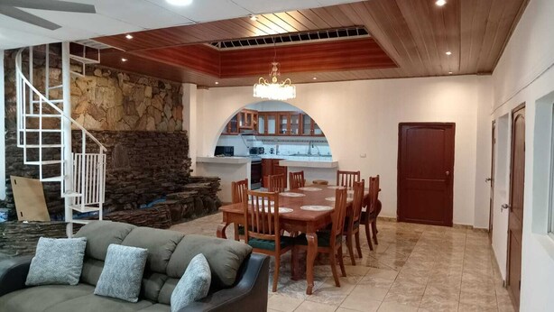 Amplia sala de estar con una mesa de comedor, techo de madera, pared de piedra y una escalera.