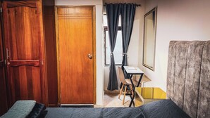Ein Zimmer mit Holztür, einem kleinen Tisch mit Stuhl, einem Bett und einem großen Spiegel an der Wand.