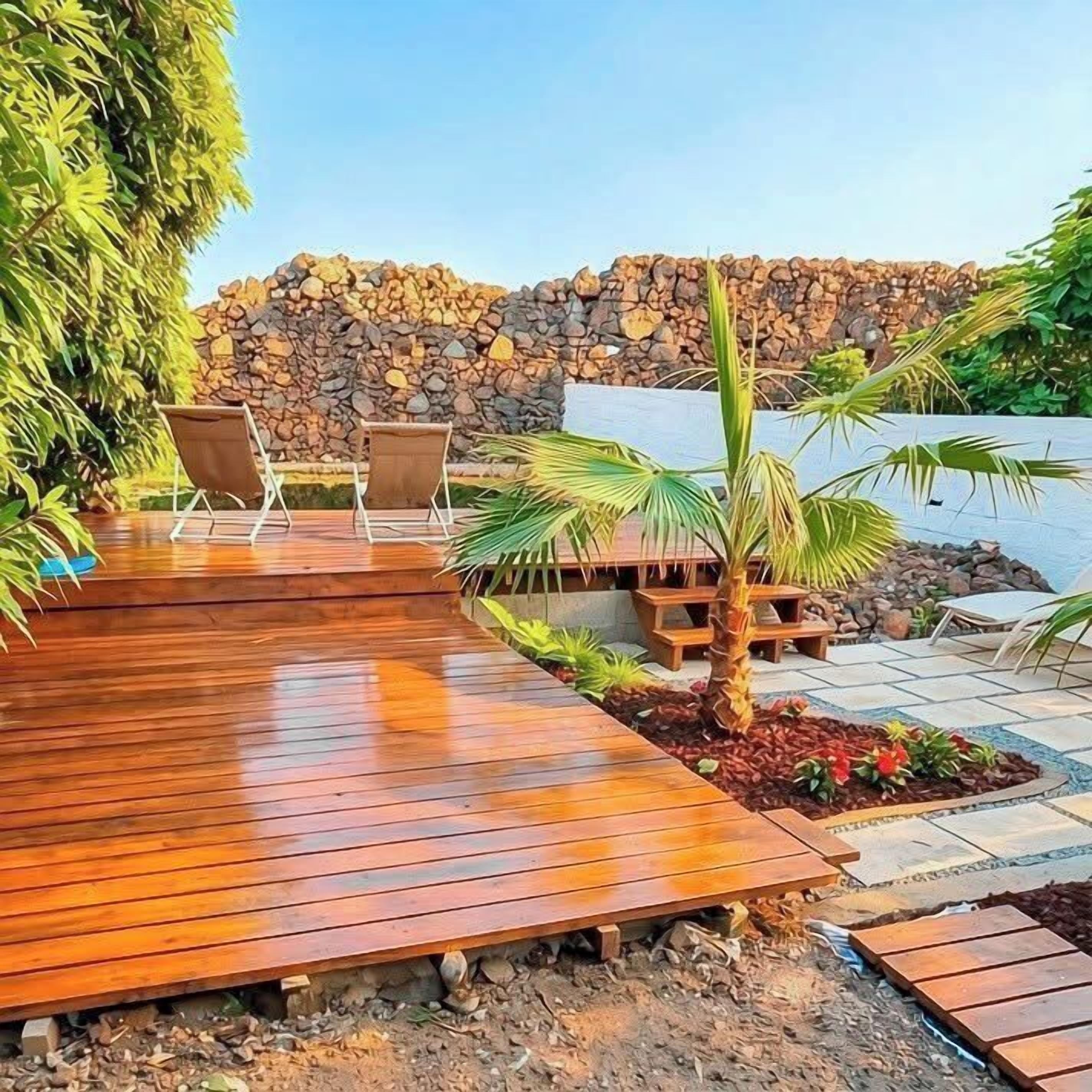 Una terraza de madera con sillones de descanso, una pequeña palmera y un muro de piedra al fondo.