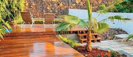A wooden deck with lounge chairs, a small palm tree, and a stone wall in the background.